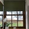 Dark green roller blind lowered over the upper half, partially blocking view and dimming light; wooden nine-pane window overlooks a grassy field with trees, sill holding white orchids and candle.