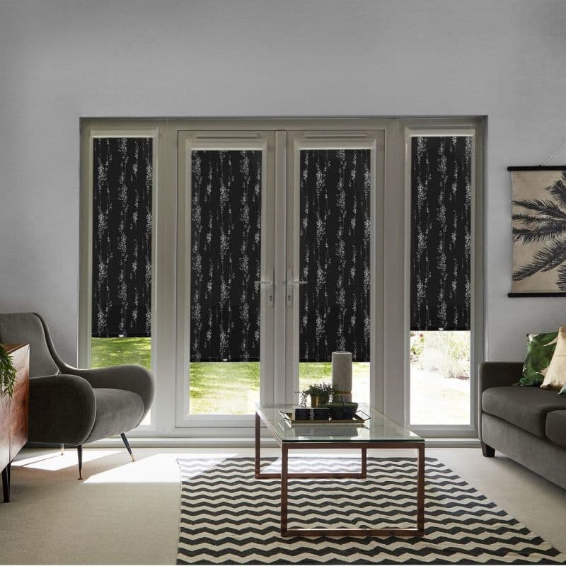 Four dark patterned roller blinds lowered over patio doors and sidelights, partially blocking sunlight; living room setting with sofas, glass coffee table, chevron rug, and sunlit lawn visible below.