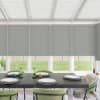 Dining table set with four place settings and candles sits beneath lowered gray roller blinds in a sunroom; green upholstered chairs surround table, windows reveal grassy backyard fence.
