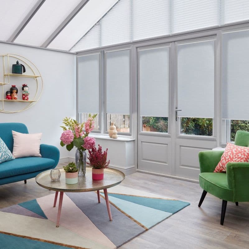Light-gray roller blinds lowered over tall glazed doors and skylights, filtering daylight; bright conservatory living room with teal sofa, green armchair, geometric rug, round coffee table and flowers; garden glimpsed.