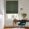 Dark green textured Roman blind, lowered with a slight bottom gap, filtering daylight over a white-framed window above a desk in a room with chair, mirror and vase of flowers.