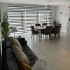 Zebra-style dual roller blinds cover a full-width floor-to-ceiling window, alternating opaque and sheer horizontal stripes filtering daylight into a bright tiled open-plan dining and living area with table and sofa.