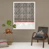 Roman-style patterned blind lowered over a white-framed window, black zigzag stripes with red trim, filtering daylight. Context: neutral-toned living-room corner with potted plant, round side table and woven chair.