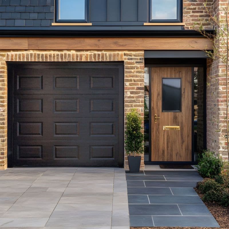 A black paneled sectional garage door and adjacent wood-paneled front door (with small rectangular glazed panel, brass handle and letterbox) are closed against a brick facade, tiled driveway, daytime light.
