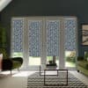French doors and sidelights are covered by navy-and-white floral roller blinds, blocking most light while revealing a lawn outside; living room contains green armchairs, glass coffee table, and chevron rug.