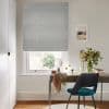 Light-blue patterned Roman blind lowered over a sash window, partially filtering daylight; sits above a wooden desk with a teal chair, mirror, vase of flowers and lit home-office interior.