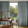Floor-to-ceiling pleated blue-gray curtains with a chevron-like pattern, parted at center; filtering soft morning light. Dining table with plates and pendant lamp in foreground, green garden visible through glass door.