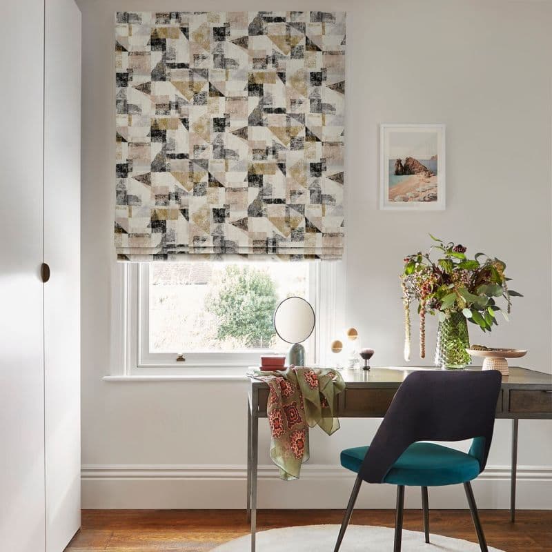 A patterned Roman blind (geometric beige, ochre, gray, black blocks) lowered over a sash window, gently filtering daylight — a bright home office desk below with teal chair and vase of flowers.
