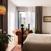 Sheer white curtains with flanking heavy grey‑brown drapes, floor‑length and partially drawn, filtering soft daylight through a glass door; bedroom contains an iron bed, tufted leather armchair, plants, rug.