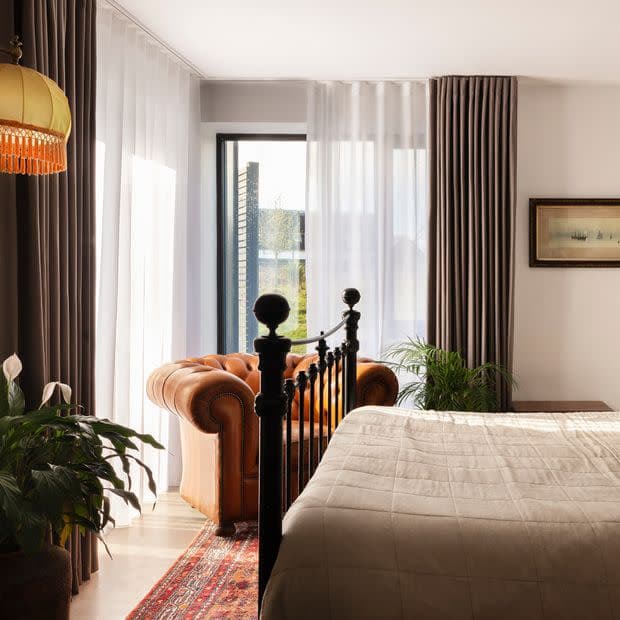 Sheer white curtains with flanking heavy grey‑brown drapes, floor‑length and partially drawn, filtering soft daylight through a glass door; bedroom contains an iron bed, tufted leather armchair, plants, rug.