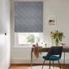 Patterned Roman blind (navy leaf print) lowered over the window, filtering daylight through a small gap; home office desk beneath with teal chair, vase of foliage, framed coastal print, walls.