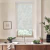 Sheer roller shade filters daylight over a kitchen window above a stainless faucet; countertop holds a bowl of greens, cutting board with cabbage, potted plants and framed sketch on wall.