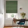 Green Roman blind, lowered with subtle folds, filtering soft daylight through a narrow bottom gap; above a desk with mirror, chair and vase of flowers in a bright home office.