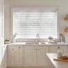White horizontal blinds covering a large window, lowered with slats mostly closed, filtering soft daylight; above a farmhouse-style sink in a bright, neutral kitchen with countertops and utensils.