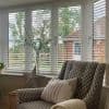White horizontal Venetian blinds over a bay window, slats tilted partially open filtering daylight and framing a garden view; cozy living-room corner with patterned armchair, glass vase and metal star ornament.