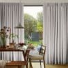 Pleated light-gray curtains, full-length and hung high, parted centrally—partially drawn and filtering daylight—framing a breakfast dining table with pancakes and a sunlit garden beyond with trees and a shed.