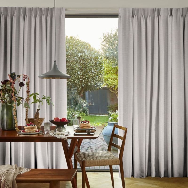 Pleated light-gray curtains, full-length and hung high, parted centrally—partially drawn and filtering daylight—framing a breakfast dining table with pancakes and a sunlit garden beyond with trees and a shed.