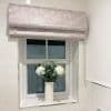 Light-pink Roman blind, folded with a bead-chain at the side; partially lowered over the top pane. Tiled bathroom, frosted window and ceramic vase with white flowers on the sill.