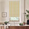 A colorful vertically striped roller blind covers a window above a kitchen sink, framed by a faucet, countertop with bowls of greens, potted plants, and framed sketch on the wall.