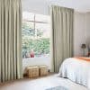 Pale green patterned pleated curtains, floor-to-ceiling and partially drawn, framing a wide sash window and filtering soft daylight; bedroom interior with bed, wicker baskets and leafy garden outside.