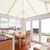 Cream pleated (cellular) blinds covering the pitched conservatory roof and windows, closed and diffusing bright daylight; small dining area with wooden table, upholstered chairs, kitchen sink and patio view.