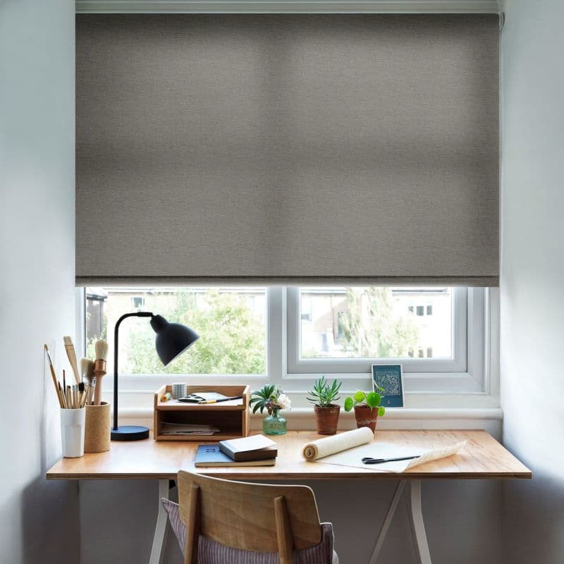 Grey fabric roller blind, lowered and dimming light through a faint cross-shaped shadow; set above a wooden desk with lamp, notebooks, potted succulents, and crafts in a bright home office.