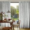 Floor-length pleated curtains patterned with pale leaves, pulled aside to frame a central opening over sliding doors; revealing a sunlit garden beyond and a breakfast-laid wooden dining table.