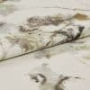 A folded cream fabric with watercolor-style olive, brown, and gray splotches; the folded edge lies across another layer, revealing a textured weave in a lit close-up on a flat surface.