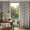 Pleated geometric-patterned curtains, drawn to either side, framing a central glass door; they partially filter light while revealing a sunlit backyard, with a dining table set for breakfast in the foreground.