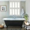 White plantation shutters (six-panel, horizontal slats) cover the central window, filtering bright daylight into a pale paneled bathroom with a black clawfoot tub, towel, plant, and bench.