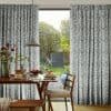 Full-length pleated curtains with blue floral print, pulled aside to frame an open gap; filtering daylight into a dining area with wooden table set for breakfast and a backyard.