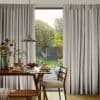 Pleated, floor-length grey curtains drawn partly open to frame a central glass door, filtering soft daylight. Dining table with breakfast plates and pendant lamp overlooks a leafy backyard garden.