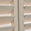 Light unfinished wood plantation shutters with angled horizontal louvers and a central stile — louvers tilted open, filtering soft daylight — close-up interior view of adjacent panels.