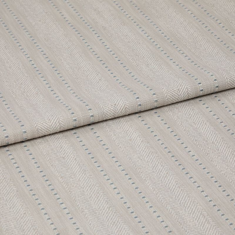 Beige woven fabric featuring herringbone chevrons and thin blue dotted vertical stripes — folded over itself, displayed as a close-up textile sample on a neutral, softly lit surface.