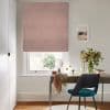 Pink textured Roman blind lowered over sash window, gently filtering daylight; light home office with wooden desk, teal chair, round mirror, vase of flowers, and garden visible beyond.