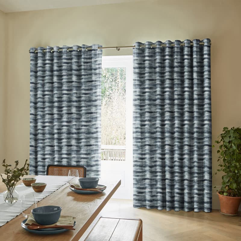 Blue-gray, horizontally striped grommet-top curtains hung on a metal rod, mostly closed with a central gap revealing a glass door; sunlit dining room with wooden table, bench, and potted plant.