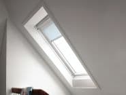 Light-blue roller blind, mounted inside an angled skylight, partially lowered and filtering daylight; set in a white sloped attic ceiling above a wooden headboard with draped cloth.