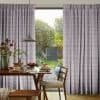 Floor-length pleated patterned curtains, pale grey with pink flecks, partially parted to frame a sliding glass door, filtering soft daylight over a breakfast table and lush garden view.