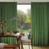 Green pleated floor-length curtains pulled aside centrally, textured fabric framing a sunlit backyard with trees and shed; a dining table with pancakes and a pendant lamp fills the foreground.