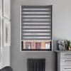 Gray zebra roller blind, lowered with alternating opaque and sheer horizontal bands, filtering light and partially revealing a brick building through the lower pane; kitchen corner with espresso machine, radiator.