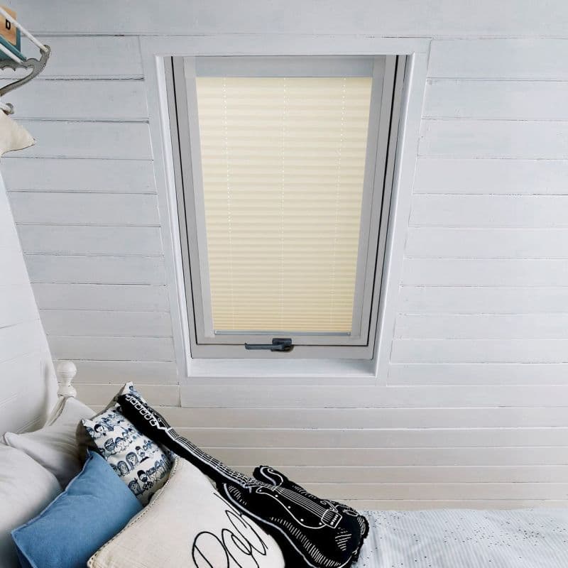 Cream pleated skylight blind, fully lowered with visible dotted ladder tapes, softly filtering daylight through a roof window; set in a white-painted attic bedroom with horizontal wood paneling and pillows.