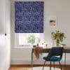 Patterned navy Roman blind covering the window, with white abstract looped motifs; lowered, filtering daylight; in a bright office with wooden desk, teal chair, vase of flowers and seaside print.