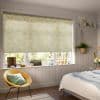 A yellow-floral roller blind lowered over a multi-pane window, filtering soft daylight into a cozy paneled bedroom with a wicker chair, hanging plant, windowsill ornaments and a bed.