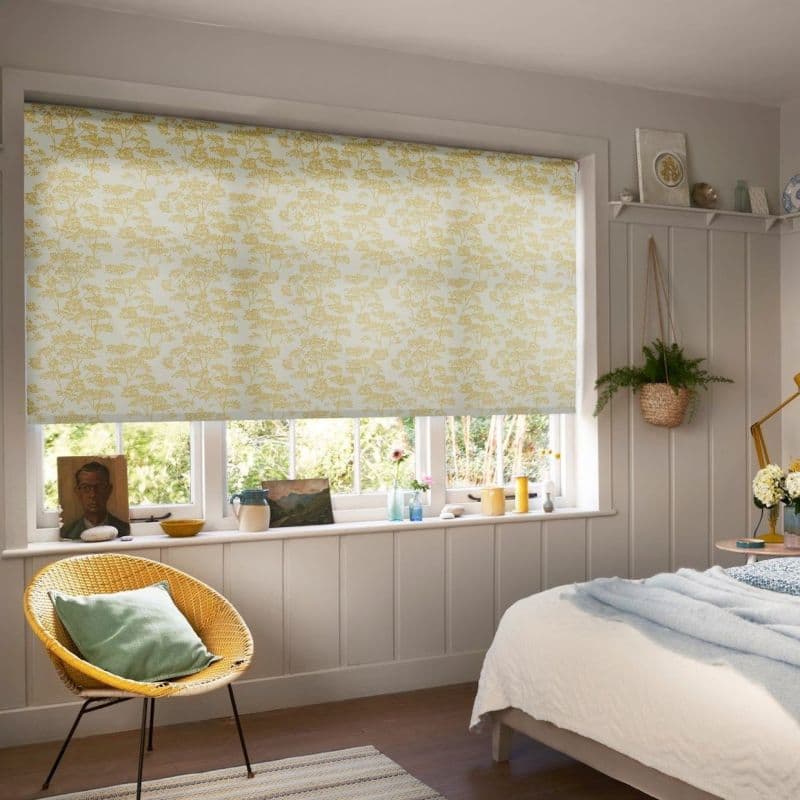 A yellow-floral roller blind lowered over a multi-pane window, filtering soft daylight into a cozy paneled bedroom with a wicker chair, hanging plant, windowsill ornaments and a bed.
