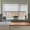 Patterned roller blind with green leaf motif, lowered across the window, filtering daylight over a kitchen sink and wooden counter; potted plant on the sill, knife block right, fence outside.