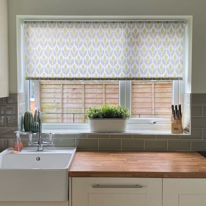 Patterned roller blind with green leaf motif, lowered across the window, filtering daylight over a kitchen sink and wooden counter; potted plant on the sill, knife block right, fence outside.