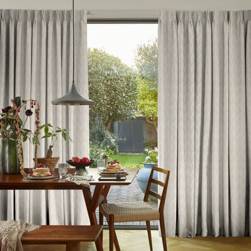 Pleated beige floor-to-ceiling curtains, drawn partly aside to frame the doorway, filtering soft daylight into a dining nook with wooden table, pendant lamp, breakfast plates and a leafy backyard.