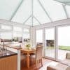 White pleated blinds covering the pitched conservatory roof and glazing, closed and diffusing bright daylight; conservatory dining area with wooden table, upholstered chairs, kitchen sink, and patio garden view.