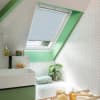 A white pleated skylight blind, lowered and filtering daylight, covers a sloped window in a bright attic bathroom with green trim, white herringbone tiles, sink, bath, plant and toiletries.