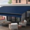 Navy blue retractable patio awning, fully extended with black roller bar, shading a glass-door terrace; outdoor seating, wooden coffee table and cushions on a sunlit deck beside a brick wall.